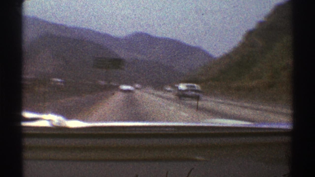 Windshield view of cars on a mountain highway with hills and distant mountains ahead.