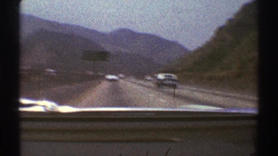 1969 Los Angeles-area 8mm windshield view of highway traffic moving through rugged hills and distant mountains.