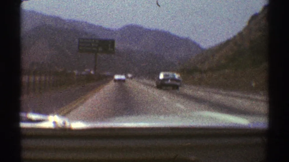 Windshield view from a 1969 home movie showing traffic, a roadside sign, and mountain slopes near Los Angeles.