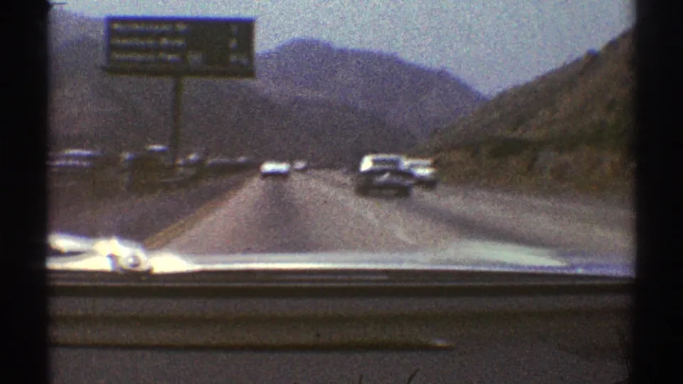 1969 8mm windshield shot of a mountain highway with a large sign, period cars, and distant ridgelines.