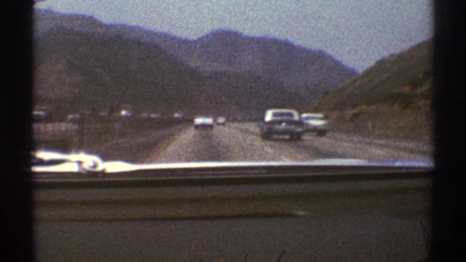 Windshield view of 1969 Southern California highway travel, with multiple cars, mountain slopes, and visible film grain.