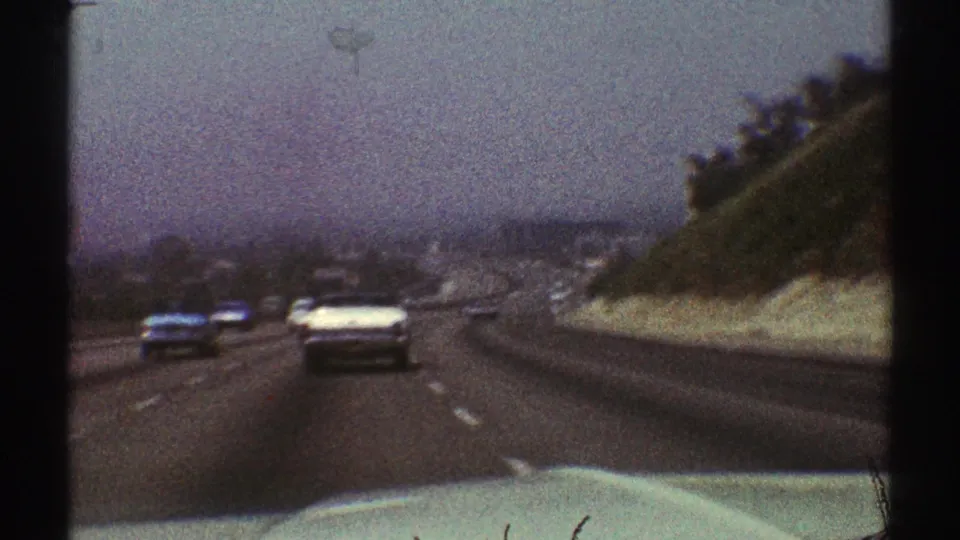 Classic cars move along a Los Angeles highway in 1969, with a sloped embankment to the right and hazy city forms in the distance.