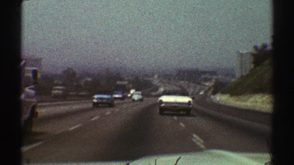 Passenger-view 8mm frame of 1969 Los Angeles roadway traffic, showing older cars, lane markings, and distant buildings under haze.