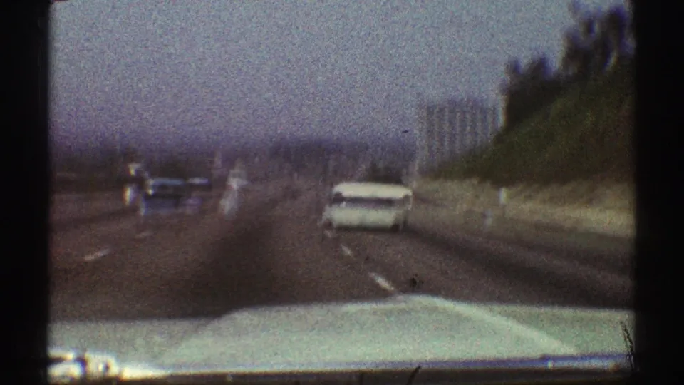 Late-1960s Los Angeles driving view with a white car ahead, roadside embankment, hazy skyline shapes, and the filming car hood at bottom.