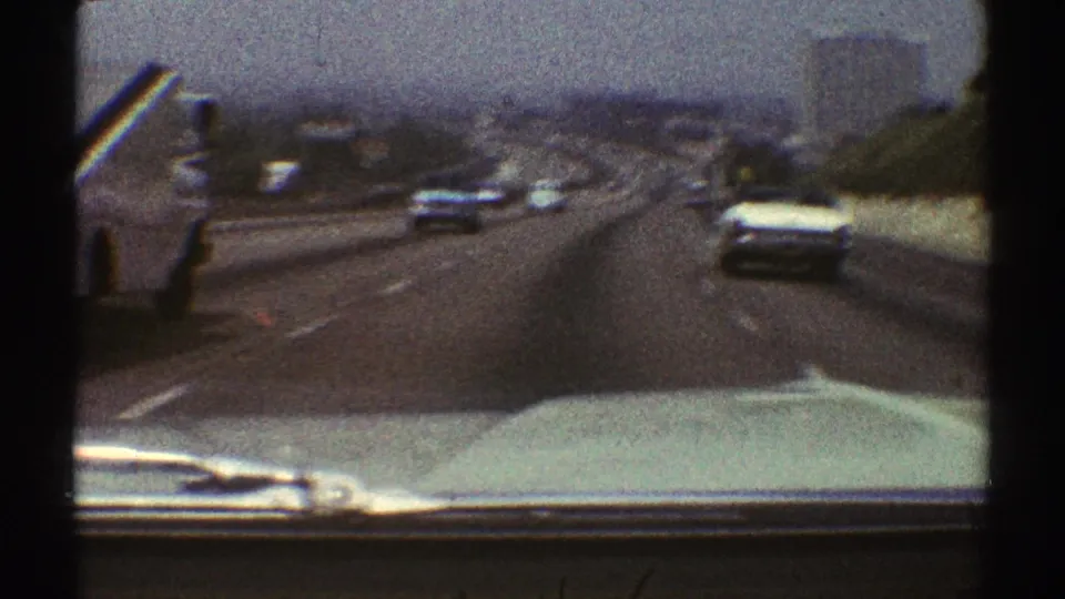 1969 Los Angeles home-movie view from a moving car on a multi-lane highway, with period vehicles ahead and a hazy city backdrop.