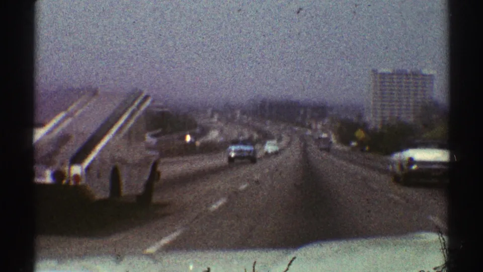 1969 Los Angeles highway view from inside or atop a moving car, with period vehicles, a distant high-rise, and faded 8mm color.