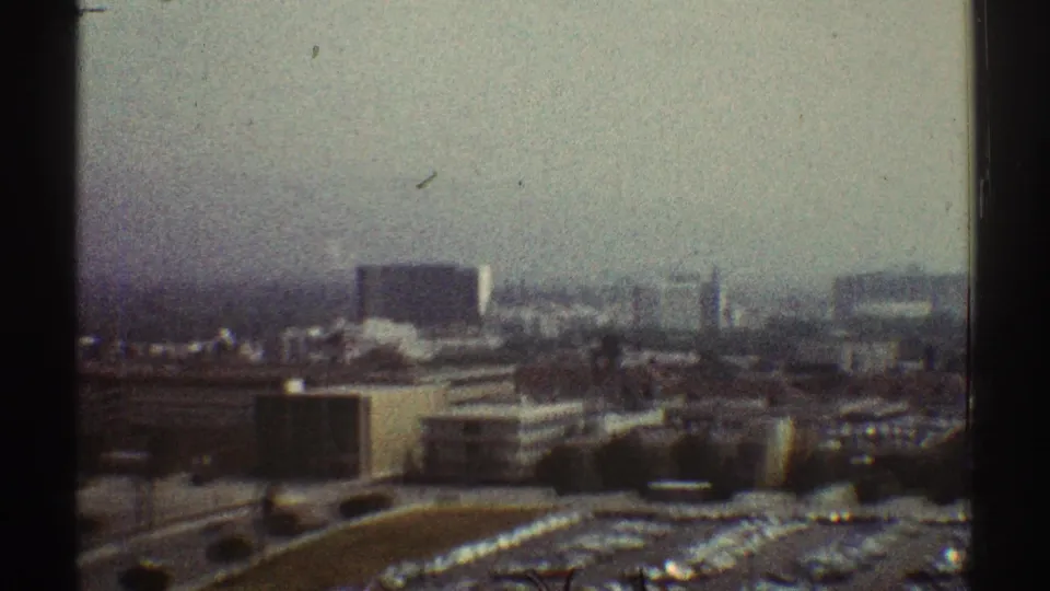 1969 Los Angeles city panorama from an elevated interior vantage, divided by a dark window frame with parking lots below.