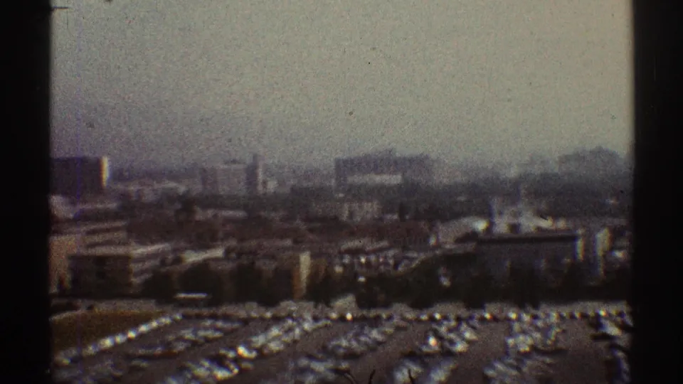 1969 Los Angeles frame with grainy haze over parking lots, rows of cars, low buildings, and distant urban structures.
