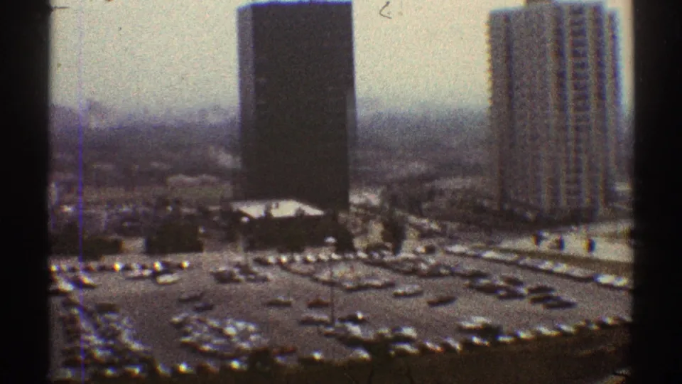 1969 Los Angeles view of a large parking area bordered by tall buildings and low structures in a soft, grainy 8mm frame.