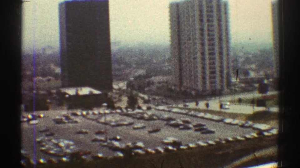 1969 Los Angeles frame showing a high-rise tower, adjacent slab-like building, and a busy surface parking lot below.