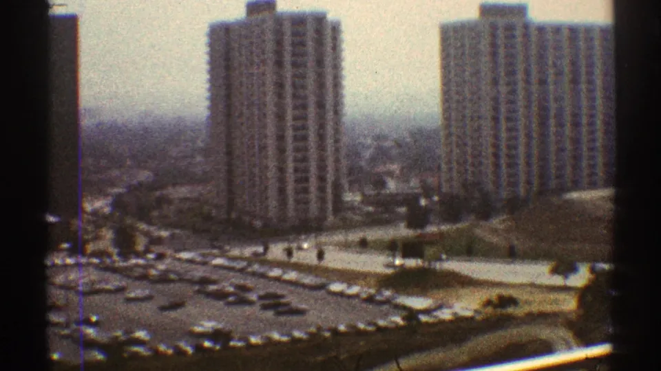 1969 Los Angeles 8mm frame with two prominent towers, a large car-filled lot, and hazy city blocks in the background.