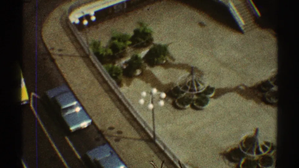 Elevated home-movie frame of a Los Angeles apartment courtyard with circular planters, lamp posts, parked cars, and an access road.