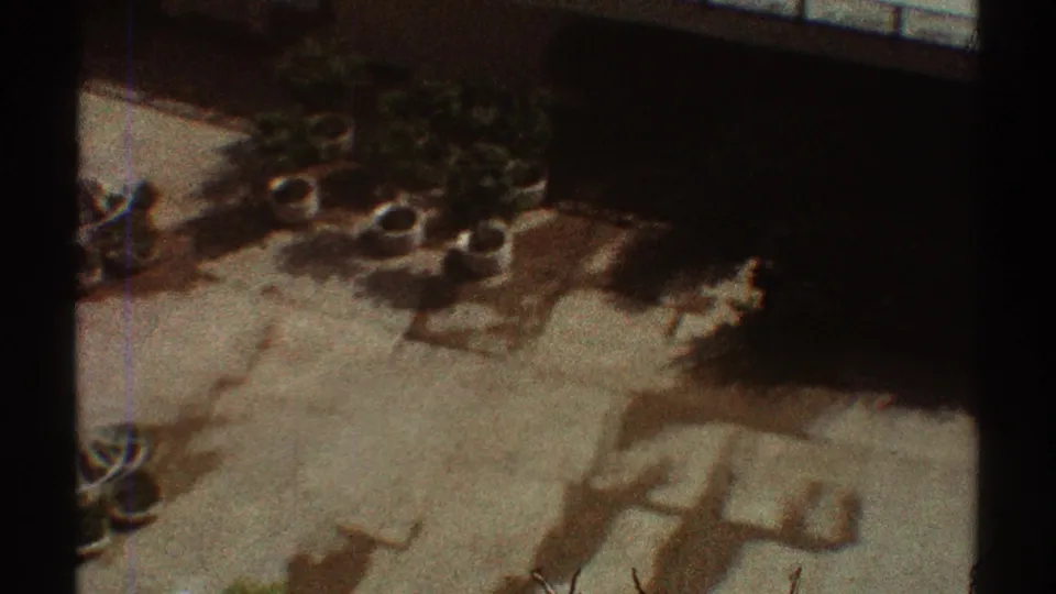 1969 Los Angeles home-movie frame looking down on a pale courtyard with circular planters, small trees, and sunlit shadows.