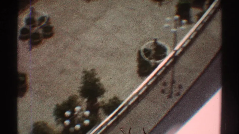 Home-movie frame of a Los Angeles courtyard corner, showing a curved border, round planters, small trees, and pale paving.