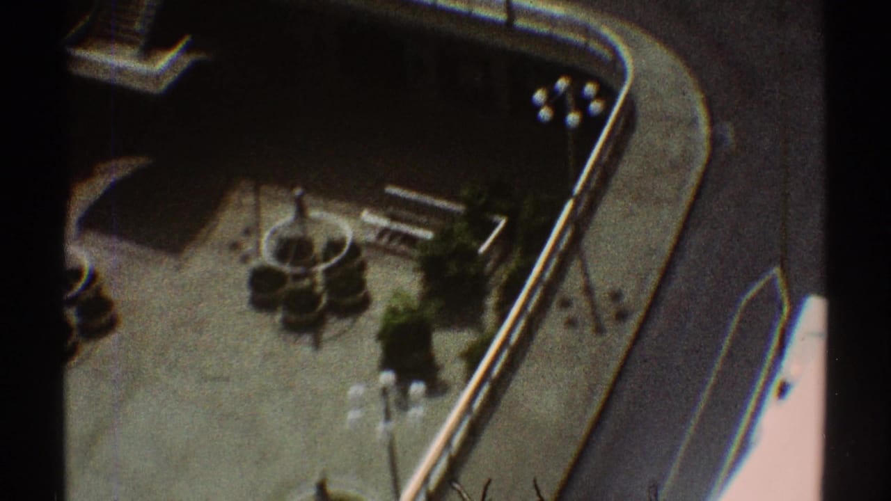 1969 Los Angeles home-movie frame showing an elevated view of a curved street edge, paved open area, landscaping, and small cars below.