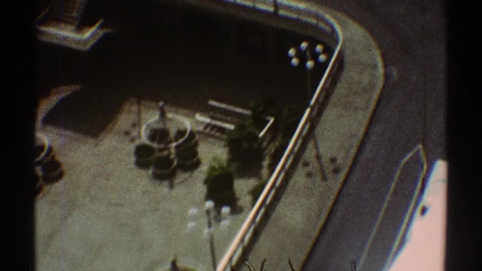1969 Los Angeles home-movie frame showing an elevated view of a curved street edge, paved open area, landscaping, and small cars below.