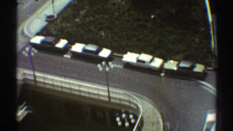 1969 Los Angeles frame looking down on parked cars, a roadway, grass, streetlights, and the corner of a modern building.