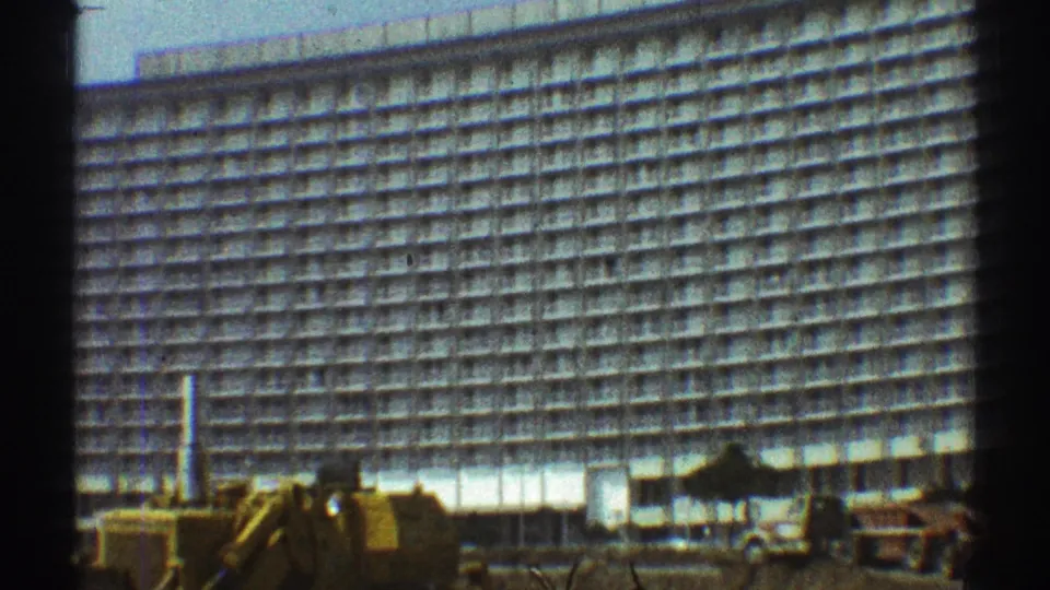 1969 Los Angeles 8mm frame showing a broad white building facade with a yellow excavator and trucks below.