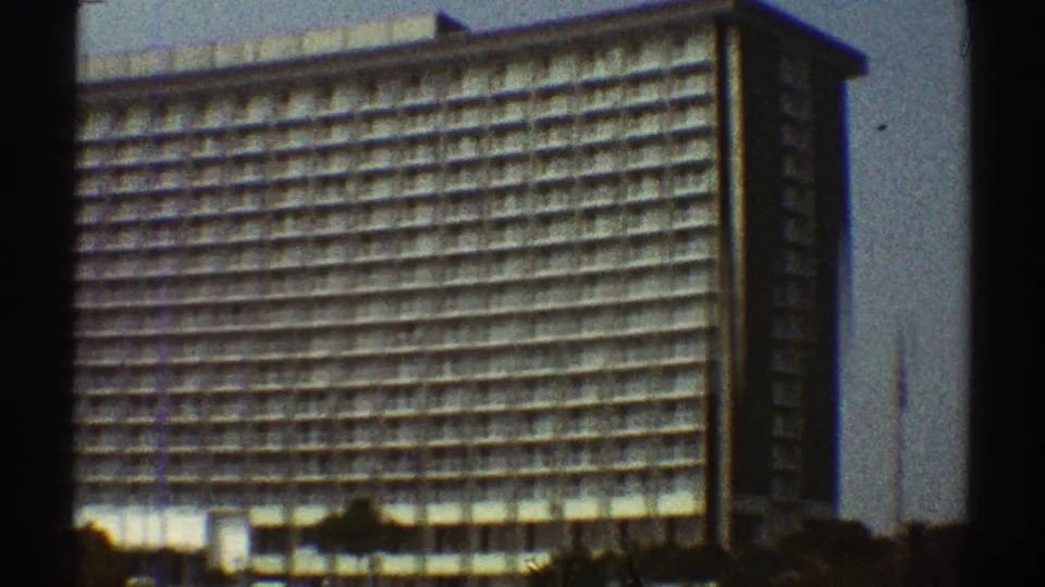 1969 Los Angeles footage frames the corner of a large hotel with stacked balcony rows, soft film grain, and low trees in front.