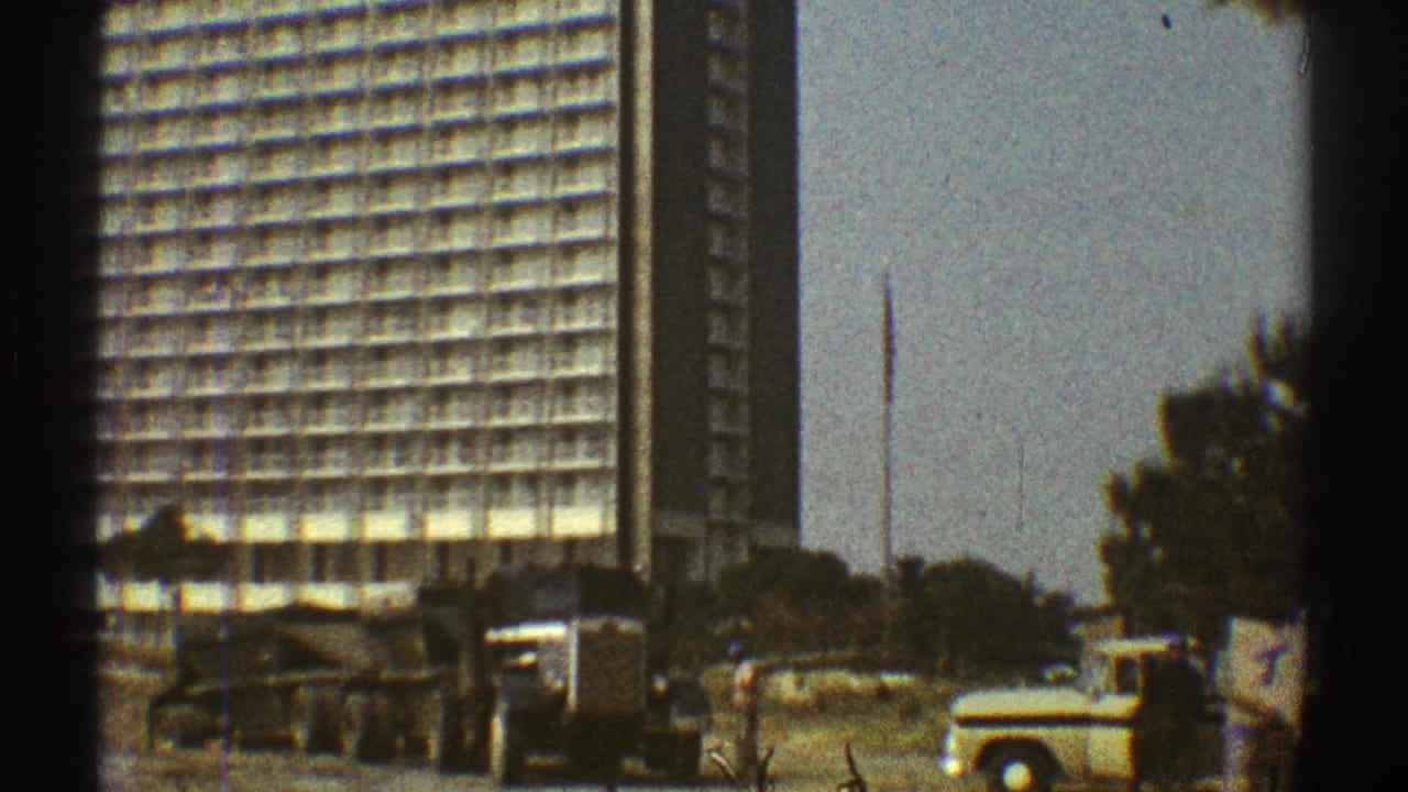 1969 Los Angeles street view with a gridded high-rise, dump trucks, roadside dirt, and a light-colored pickup or van.