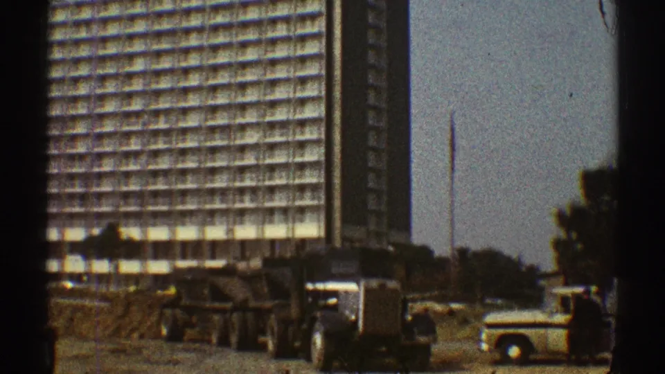 The frame shows a large dump truck crossing the foreground near a tall gridded building in 1969 Los Angeles.