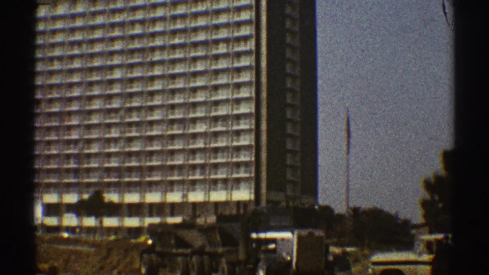 A brief 1969 Los Angeles street view captures a dump truck, a light vehicle, trees, and a gridded high-rise through 8mm film borders.