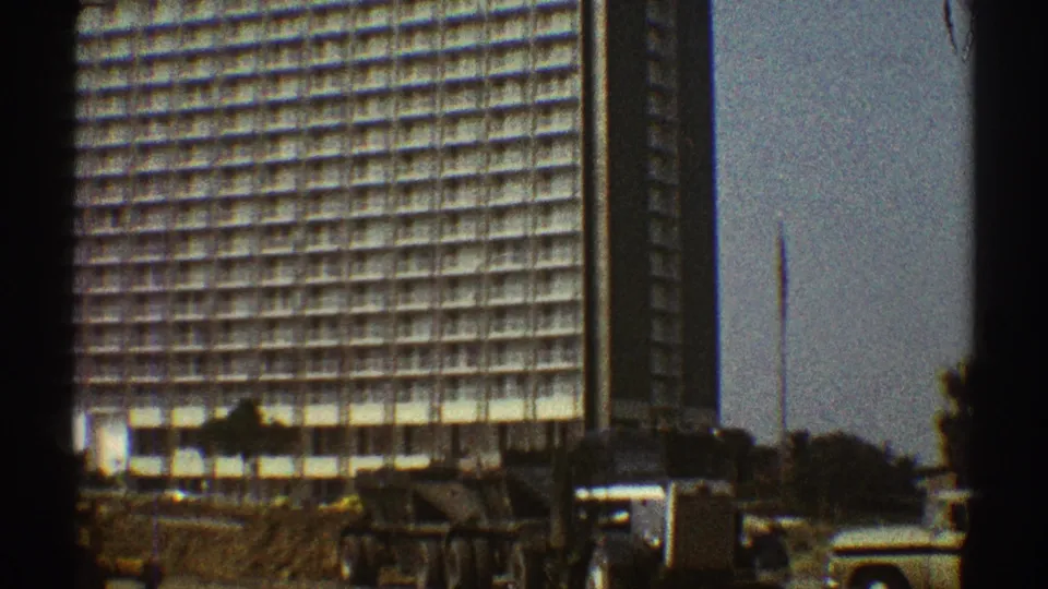 1969 Los Angeles 8mm frame showing a large gridded high-rise under construction with earth, machinery, and a pale vehicle below.