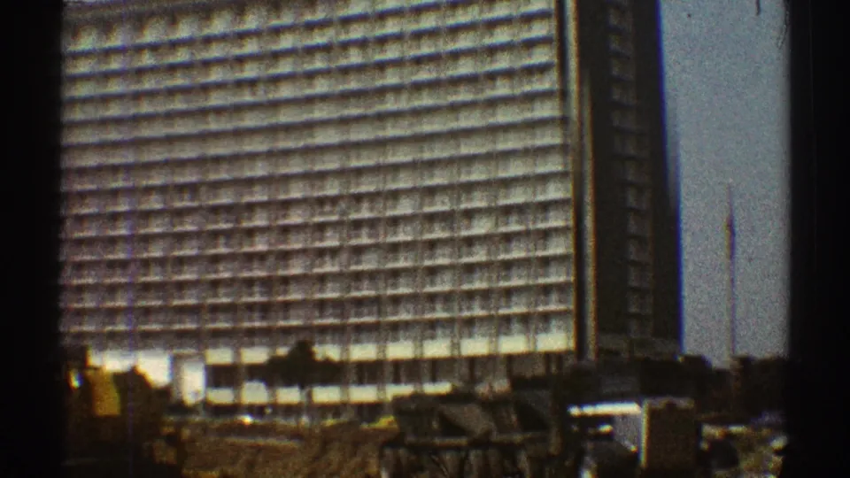 1969 Los Angeles 8mm frame with dark gate edges around a gridded high-rise, construction equipment, trees, and a flagpole.
