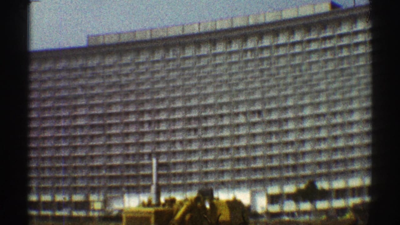 1969 Los Angeles 8mm view of a yellow tractor below a massive high-rise facade with dark film borders.