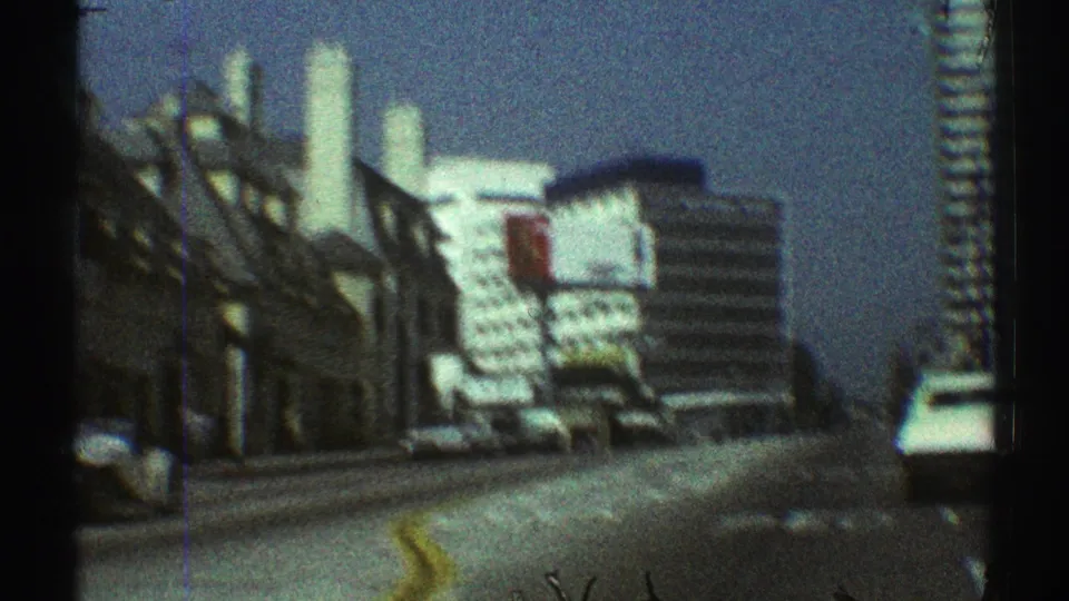 1969 Los Angeles street-level view toward white modern buildings with parked cars, a red sign, and blue sky in grainy 8mm footage.