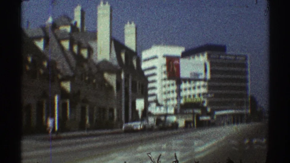 1969 Los Angeles frame showing older street-front buildings leading toward white modern high-rises and parked cars on a sunny street.
