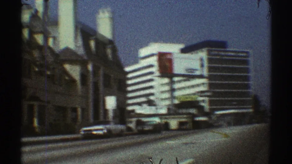 1969 Los Angeles street view of a white modern building with horizontal bands, a red sign, parked cars, and soft 8mm color grain.