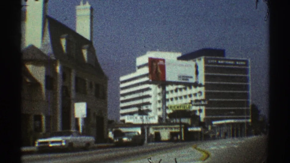 1969 Los Angeles street view from a moving car, with a white sedan passing older buildings and a striped commercial block ahead.