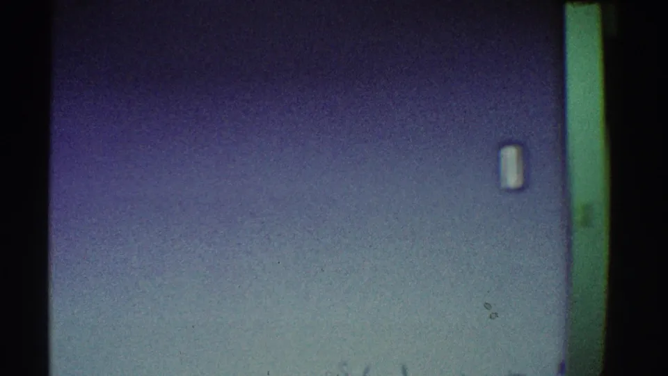 Interior wall detail showing a blue wall, small wall switch or plate, green door frame edge, and soft 8mm film grain.
