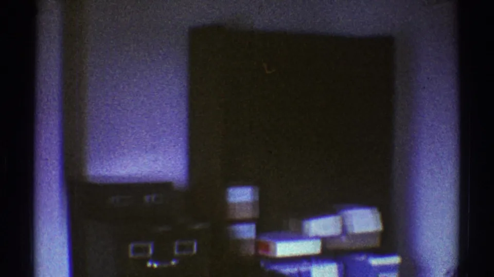 The camera reveals a cluttered Los Angeles storage room with stacked boxes, low cabinets, books, and a dark door or cabinet face.