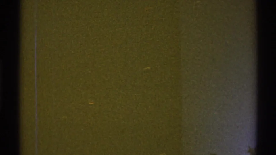 Grainy 8mm frame of a brown apartment wall and dark door frame, with muted exposure and visible film wear.