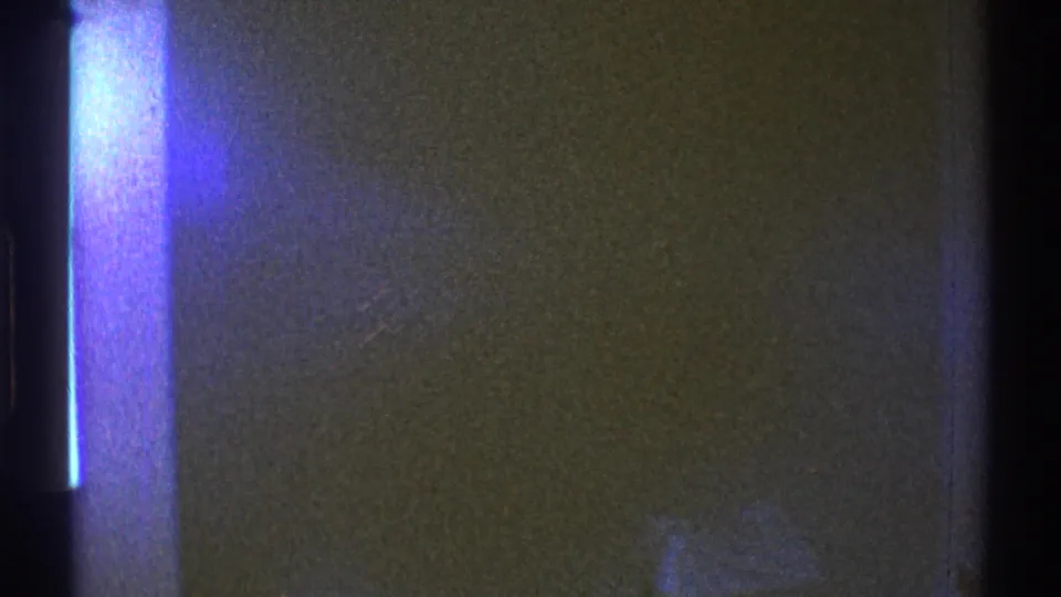 1969 Los Angeles home-movie frame with dark interior walls and a strong blue-purple light leak crossing the view.
