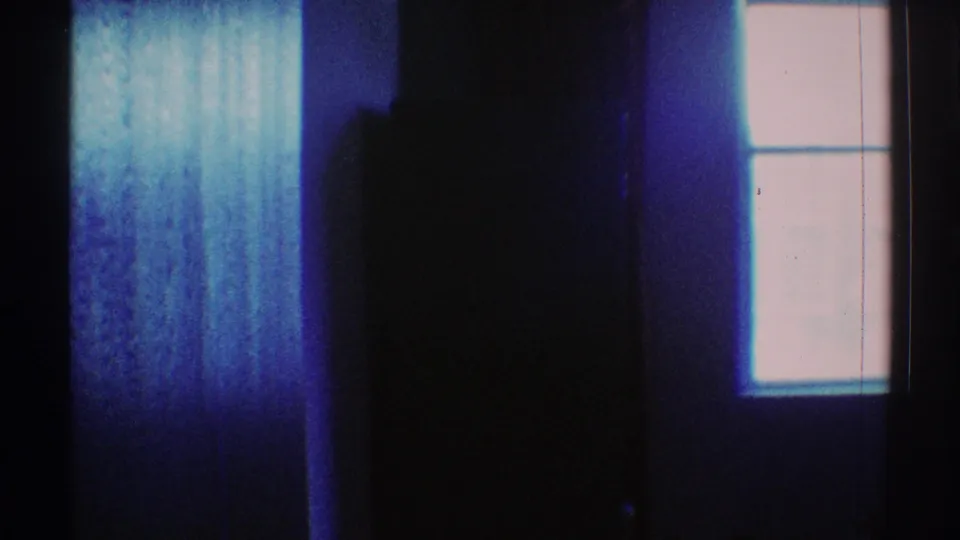 1969 Los Angeles home-movie frame showing a shadowed interior, right-side window, and blue-purple light wash.