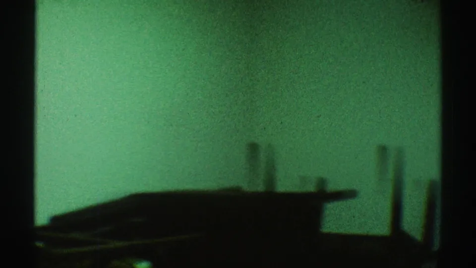 1969 Los Angeles 8mm frame with a dark furniture-like silhouette, green wall, black vertical framing, and visible film grain.