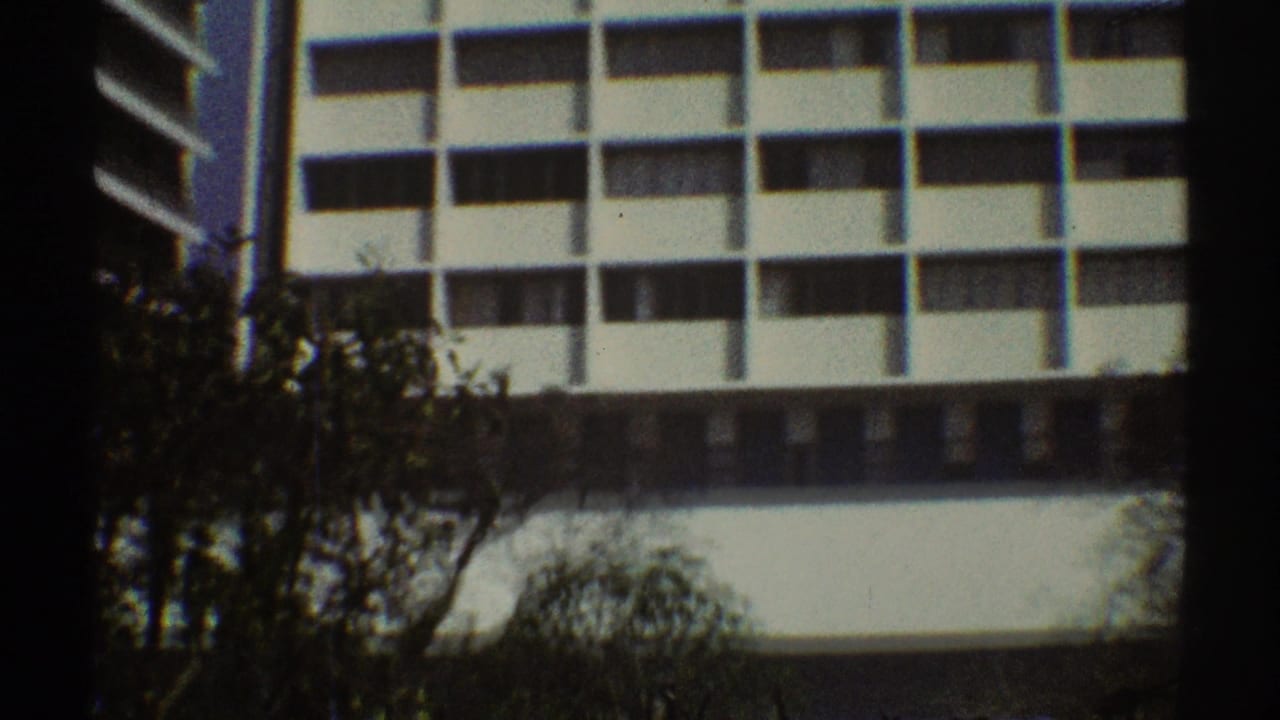 1969 Los Angeles 8mm frame showing white modern office facades, dark windows, and tree silhouettes in the foreground.