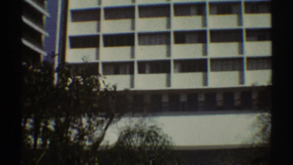 1969 Los Angeles 8mm frame showing white modern office facades, dark windows, and tree silhouettes in the foreground.