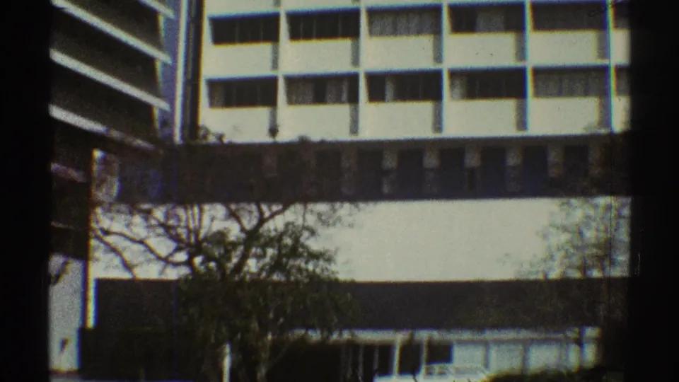 Handheld 8mm view of a white Los Angeles office building, dark window band, trees, and lower structure at street level.