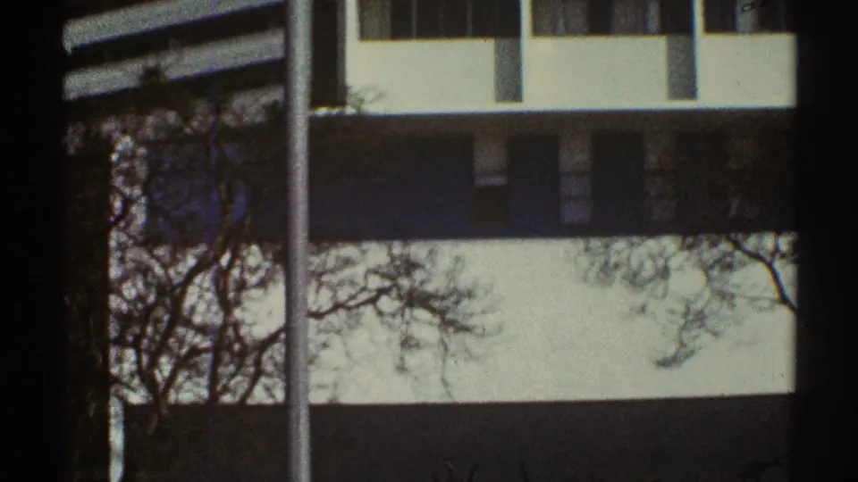 1969 Los Angeles 8mm frame of a white modernist building with dark bands, rectangular windows, bare branches, and a pole at left.