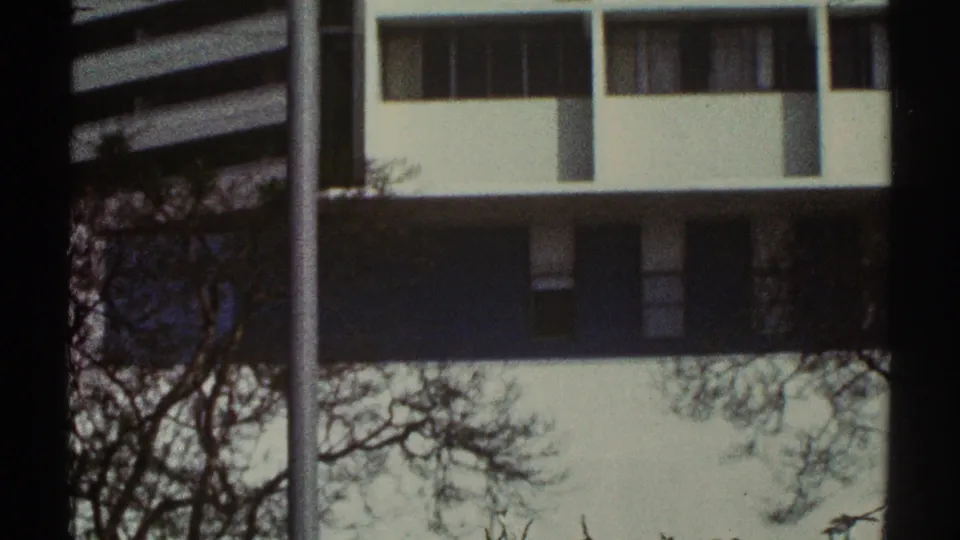 1969 Los Angeles 8mm view of a pale modernist exterior with upper windows, a dark horizontal band, bare trees, and a vertical pole.