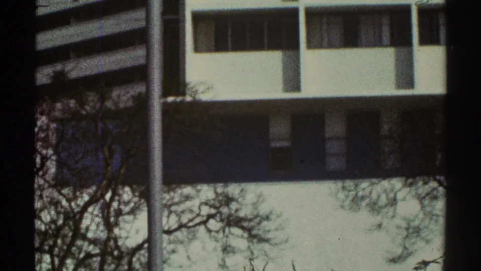 1969 Los Angeles frame showing the same white modernist building, rectangular windows, dark facade band, bare branches, and 8mm frame edges.