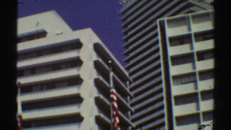 1969 Los Angeles frame with white modernist office towers, tiered balconies, American flags, and clear blue sky.