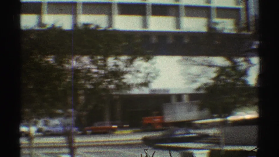 1969 Los Angeles street frame with a white overhead building section, roadside trees, traffic, and a small red vehicle.