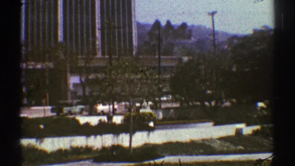1969 Los Angeles home-movie frame showing traffic, trees, a tall building, a utility pole, and hazy hills beyond the street.