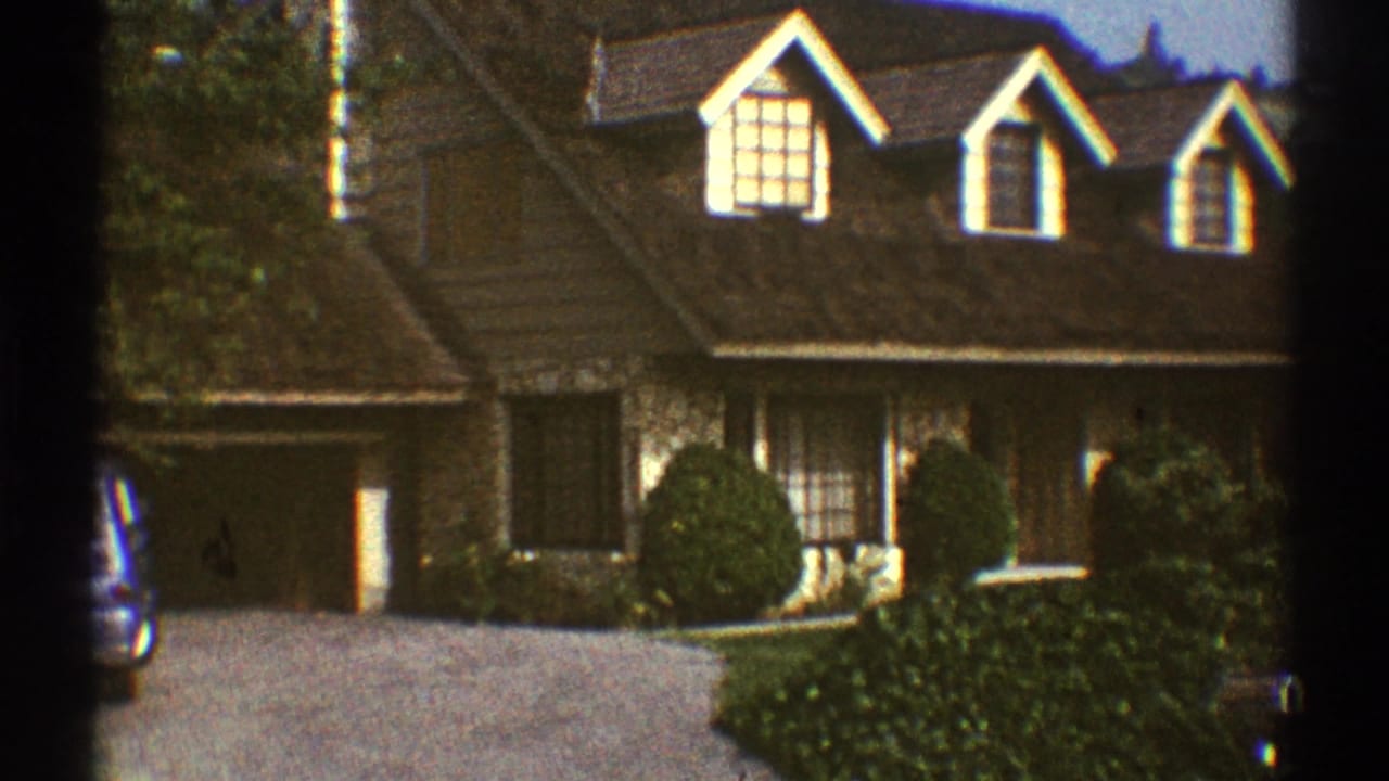 1969 Los Angeles home-movie frame showing a brown two-story suburban house with dormer windows, garage, shrubs, and driveway.