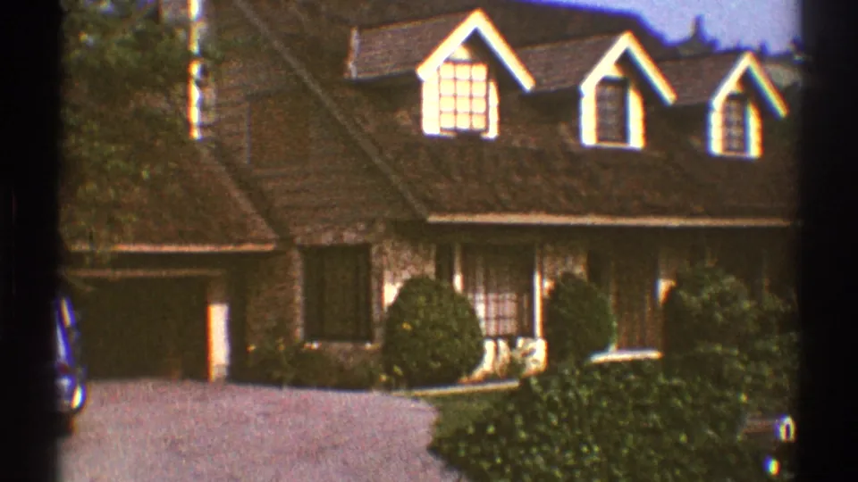 1969 Los Angeles exterior view of a large brown suburban house with three dormers, front shrubs, garage, and driveway.